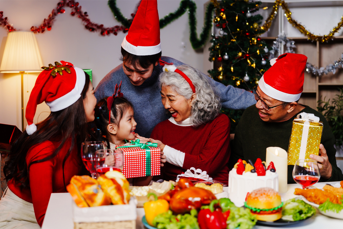 Smiling family exchanging wrapped gifts in a cozy dining room decorated with warm lights.